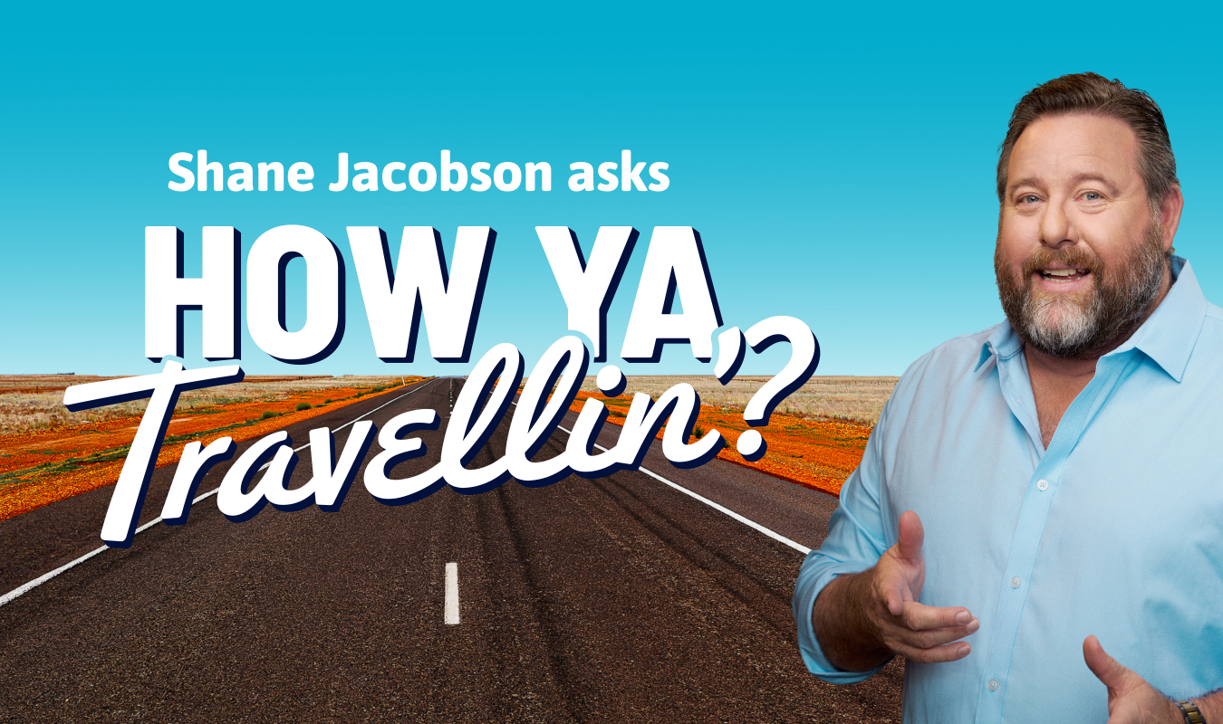 How Ya Travellin'?Campaign image that has text that reads 'Shane Jacobson asks 'How Ya Travellin'?'. To the right of the text is an image of Shane Jacobson in front of a road in the outback.